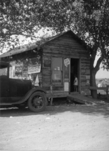 Small wooden building with ramp leading up to door and small child in doorway; old (Model T?) car outside