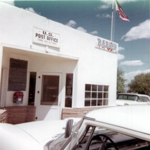 White building with American Flag, small letter box on wall, Barber sign, US Post Office sign, and cars in parking lot