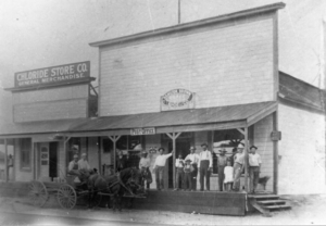 Several people standing on wooden desk outside post office building and general store; wagon with two horses in street