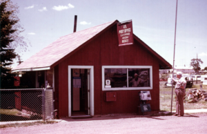 Color photo of man standing outside building with peaked roof, signage near top of building US Post Office Clay Springs, chain link fence next to building, building door open, newspaper vending machine outside