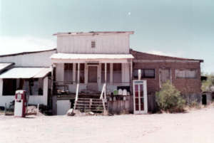 Color photo of exterior of Cleator post office building; stairs leading up to door dilapidated, gas pump, phone book, and mailboxes outside