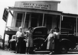 Black and white photo of four people standing by a truck with "Thomas R. Cleator" on truck door; building behingd truck is on stilts and has signage James P. Cleator Cleator Post Office