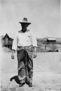 Black and white photo of man in hat standing outside several yards in front of buildings