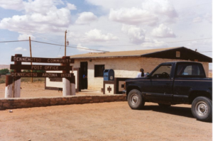 Exterior adobe building and dirt parking lot; Sign Dennehotso Community Post Office Dennehotso Ariona 85635; dark truck in parking lot, person with long black hair approaching building