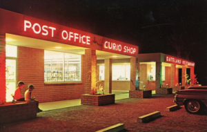 Strip mall exterior at night with neon signage Post Office Curio Shop Dateland Restaurant; two people (probably children) sitting on a planter bench; car in parking lot