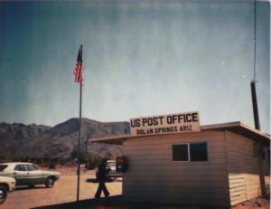 Exterior building with flat roof, small window, small letter box attached, signage on building US Post Office Dolan Springs Ariz, flag on pole, person walking towards building, dirt parking lot with three cars