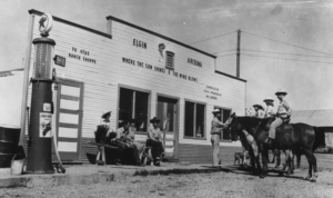 Exterior building, porch, and dirt; 4 people sitting one standing, and 3 people on horses; dog; building signage Elgin Arizona where the sun shines and the wind blows, Ye olde ranche shoppe; possible gas pump