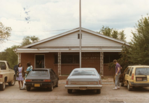 Exterior brick building with dirt parking lot, one truck and three cars, three people by one car and one person exiting another car; awning on building; post office signage on building
