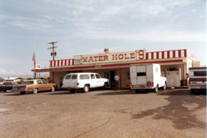 Exterior building and parking lot; top front of building has red and white stripes and signage Water Hole 9 and Post Office Fort Mojave; trucks and cars in parking lot; ice cooler on porch