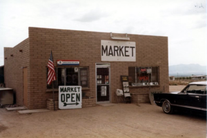 Exterior Golden Valley post office building with "Market" and "Market Open" signs, American flag, car in dirt parking lot