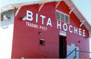 Red building exterior with "Bita Hochee Trading Post" painted; open door with stairs leading up to it