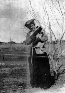 Woman with long dress and coat, wearing hat, holding small dog, outside barren ground near tree.